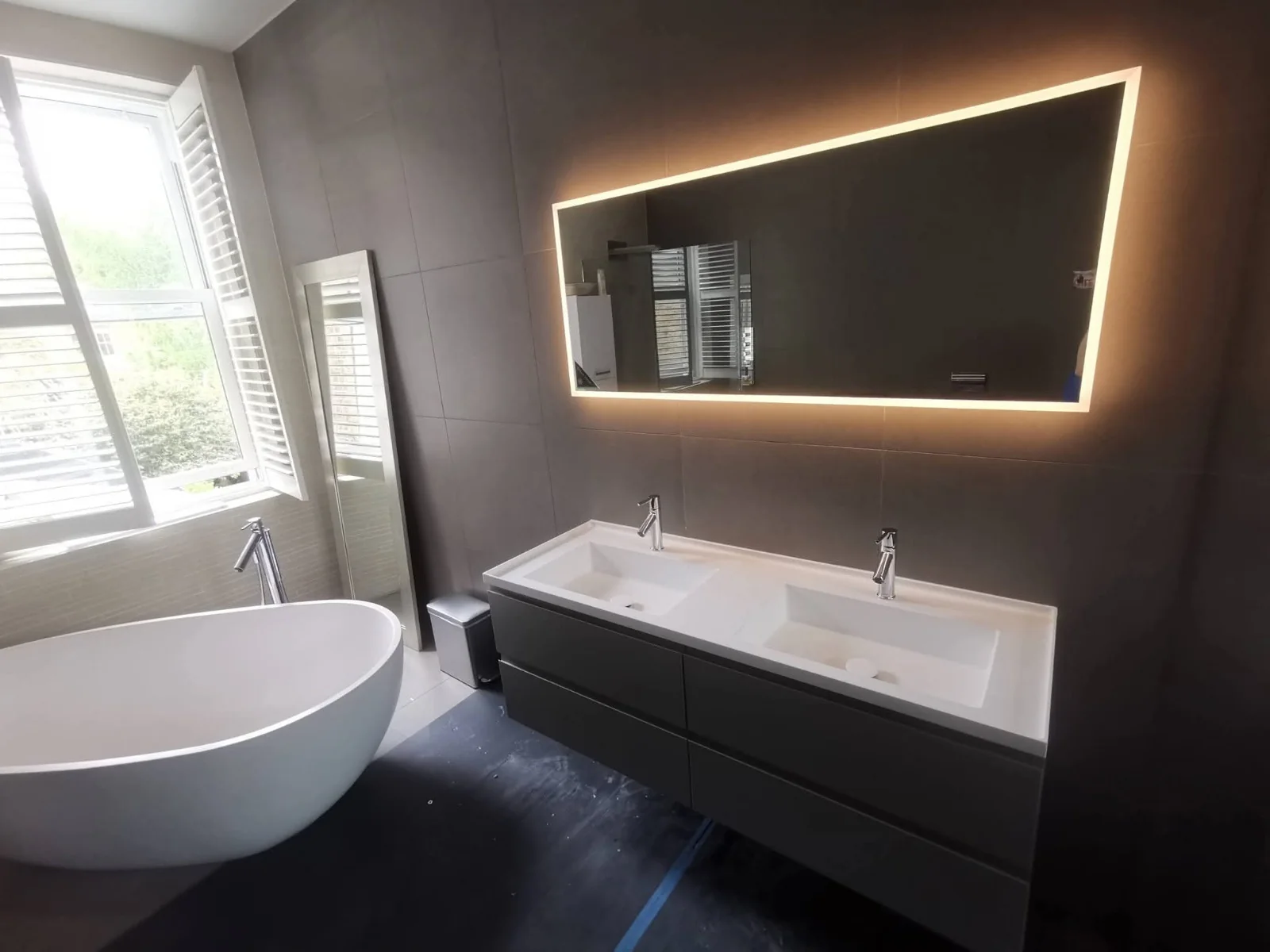 Freestanding bath and double vanity with backlit mirror in a grey-tiled bathroom by AdiDeccor